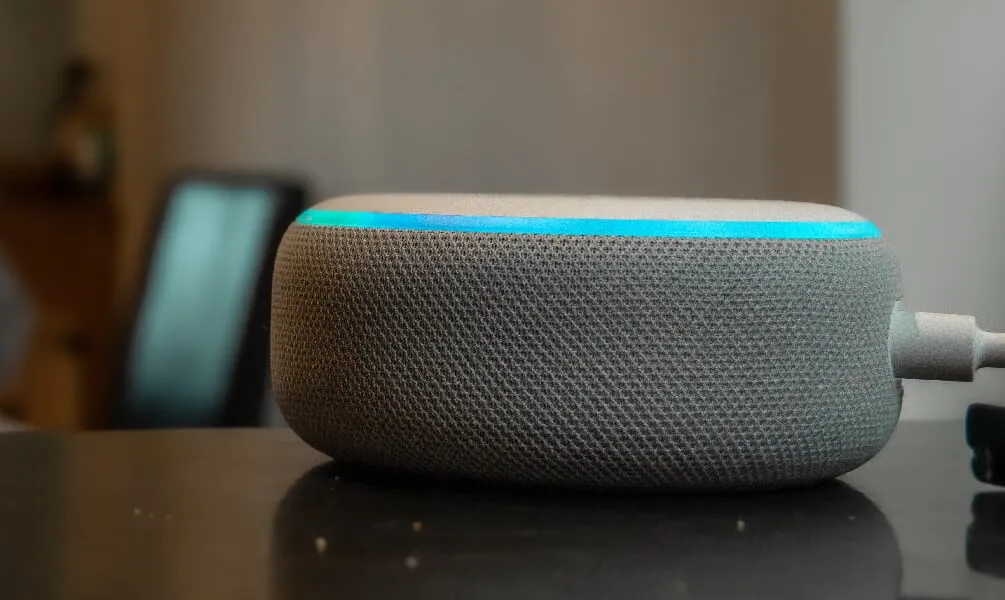 An Amazon Echo Dot smart speaker glowing with a cyan blue light on a dark reflective table.