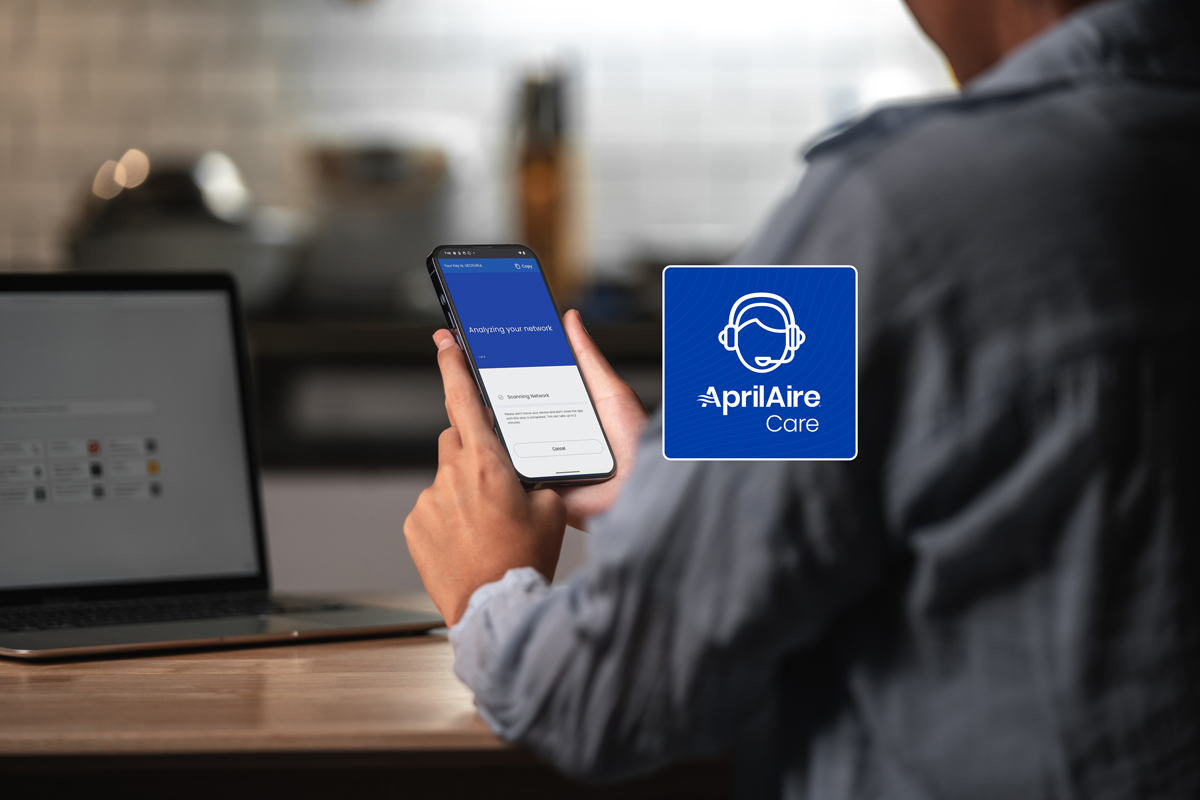 Person holding a smartphone displaying the AprilAire Care app scanning for a network, with a laptop open on the table.