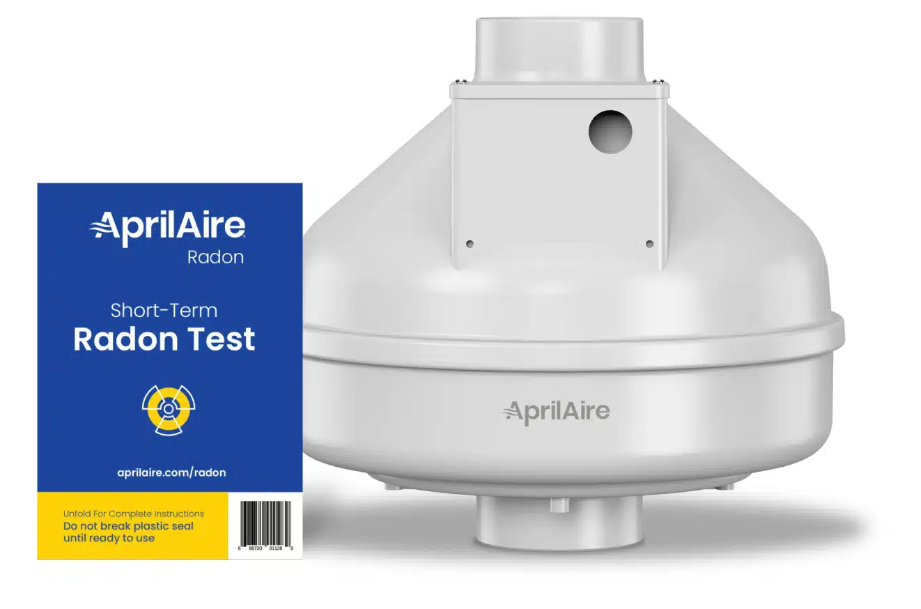 A white AprilAire radon mitigation fan unit standing next to a blue short-term radon test kit package.