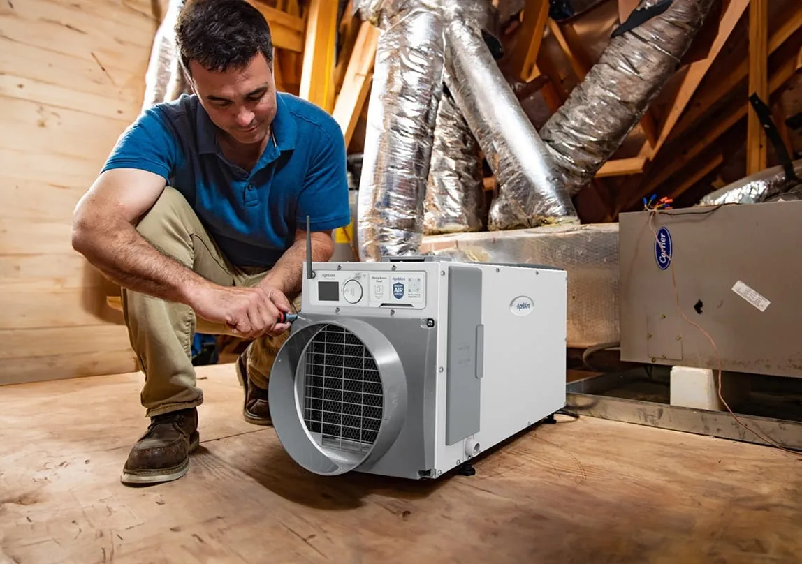 HVAC technician installing an AprilAire whole-home dehumidifier system in a residential attic next to ductwork.