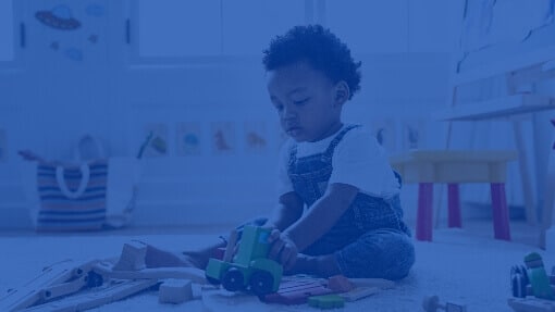 Toddler sitting on the floor in a nursery, intently focused on pushing a small green wooden train.