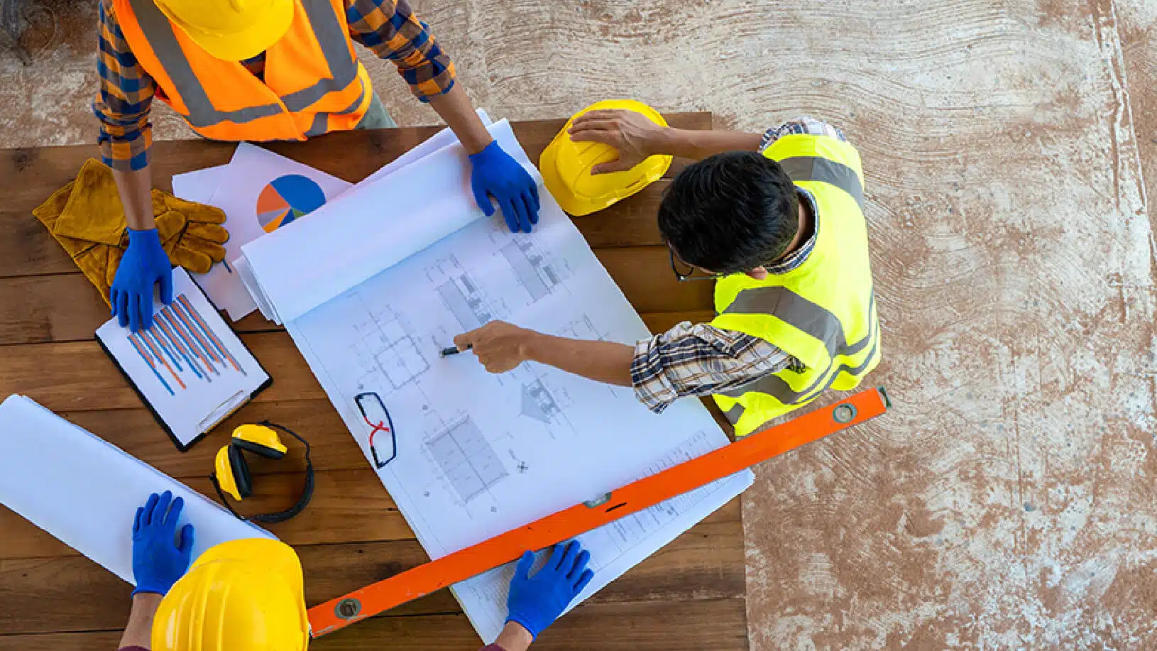 High-angle view of three construction workers in safety vests and hard hats analyzing blueprints and charts on a wooden table.
