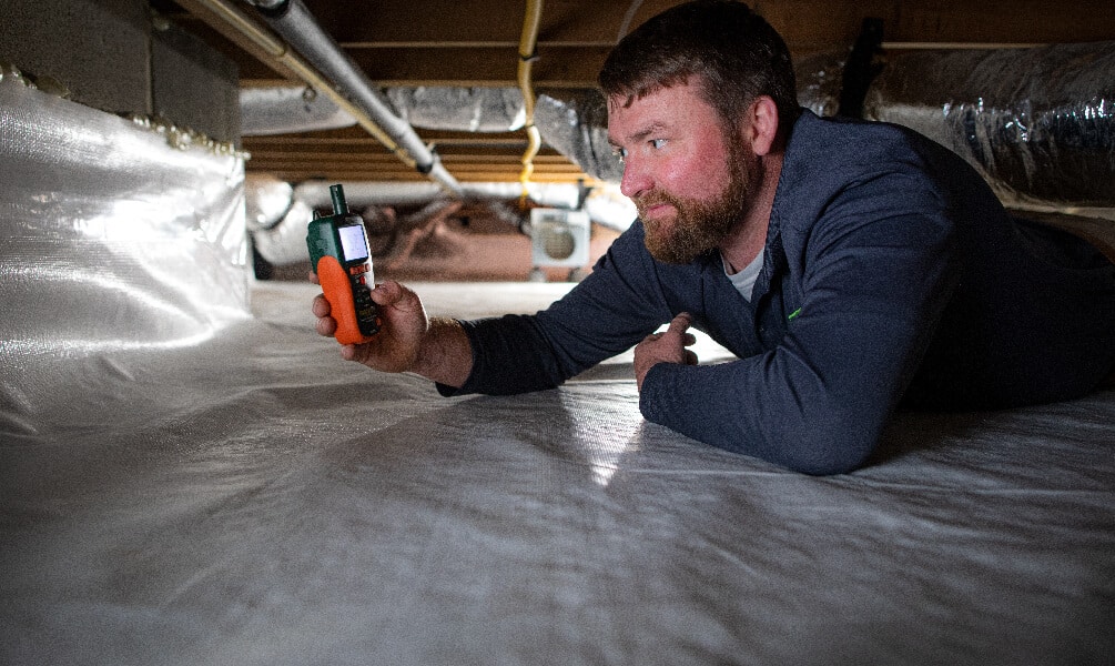 A technician crawls under a house to check moisture levels using a digital meter on a white vapor barrier.