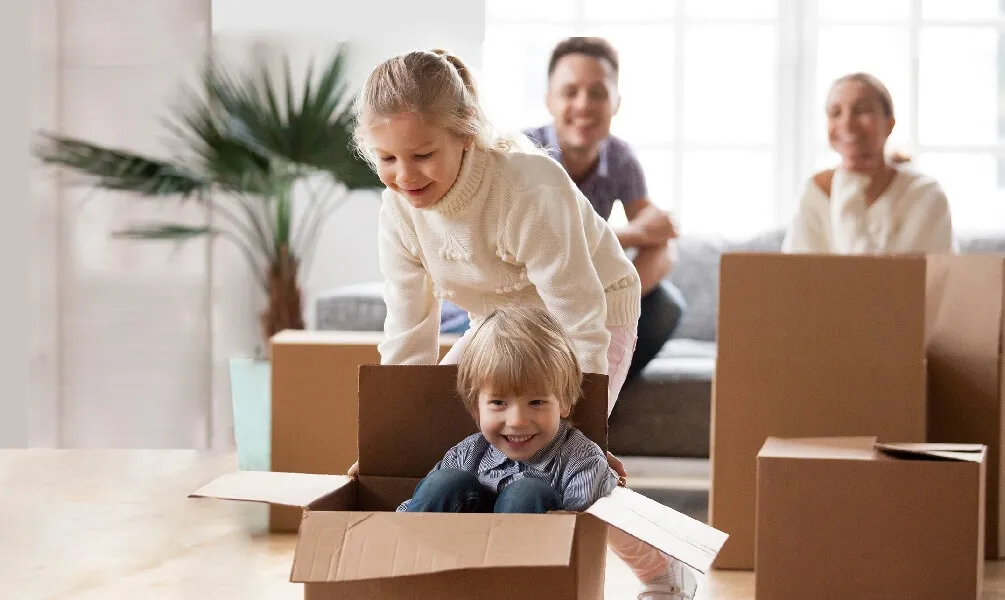 Two young siblings playing happily with cardboard moving boxes in a new home while their parents smile in the background.