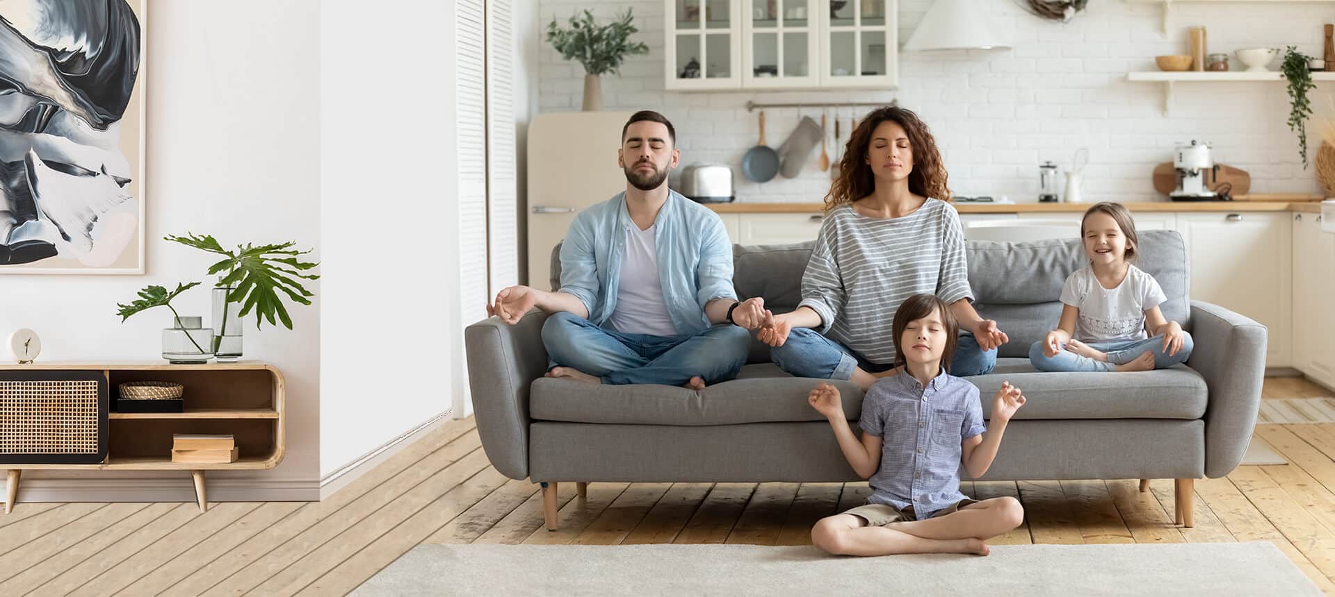 A family of four, parents and two children, sitting on a gray couch and floor in a bright home, meditating with their eyes closed.