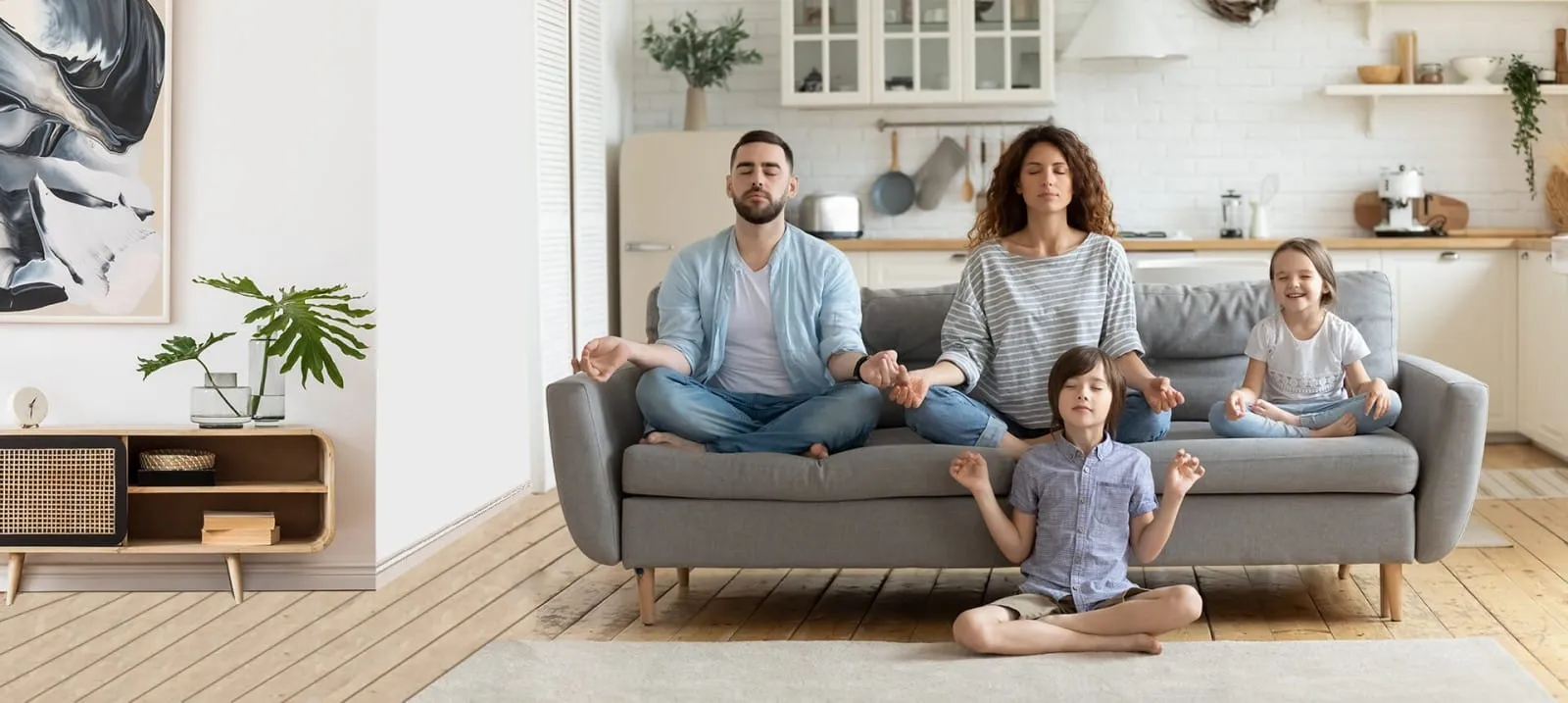 A family of four, including a mother, father, and two children, sits in a bright, modern living room meditating with eyes closed.
