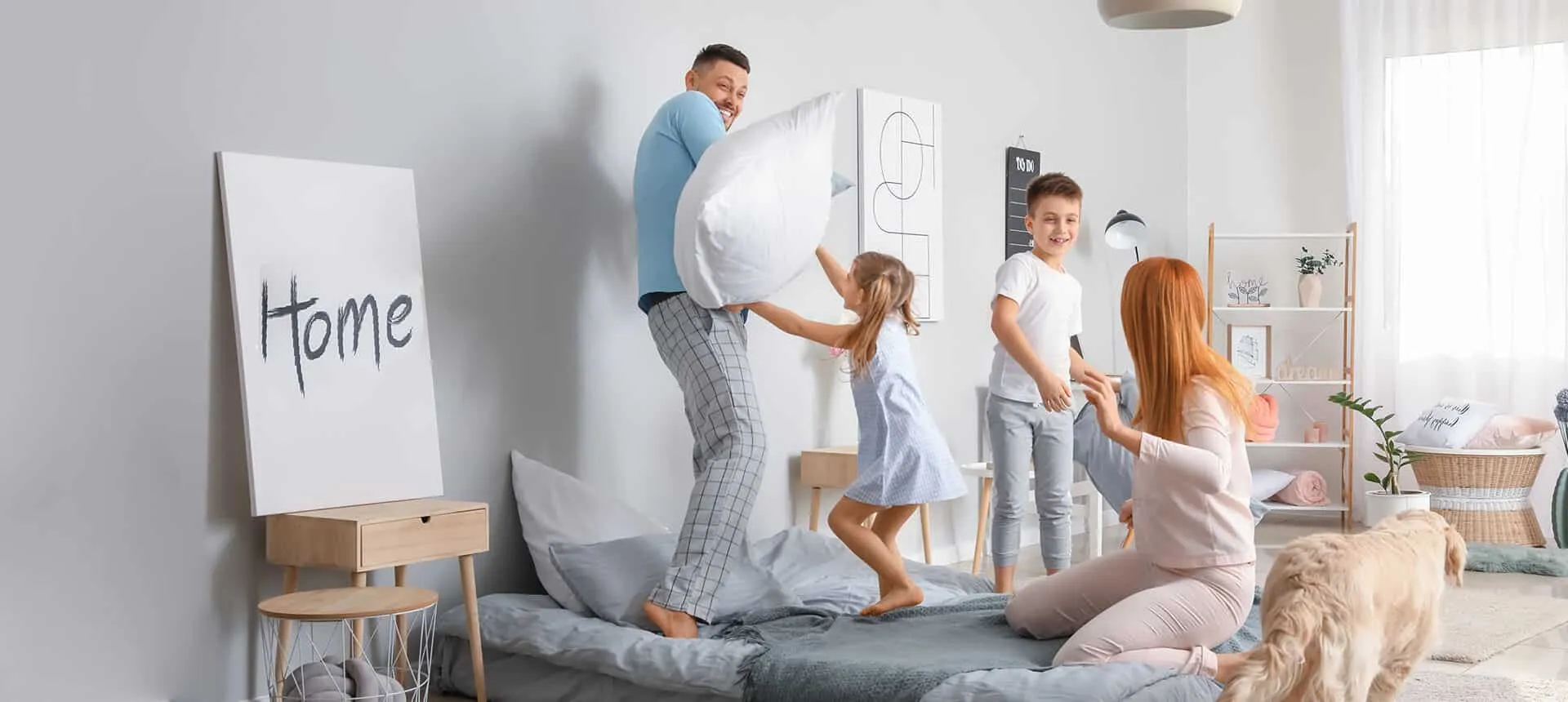 A family and their dog have a playful pillow fight in a bright, clean bedroom representing healthy indoor air