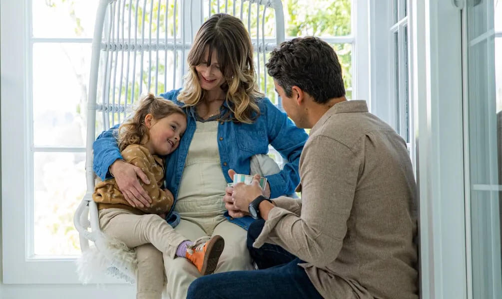 A woman sitting in a white hanging chair hugs a smiling young girl while a man sits nearby holding a mug.