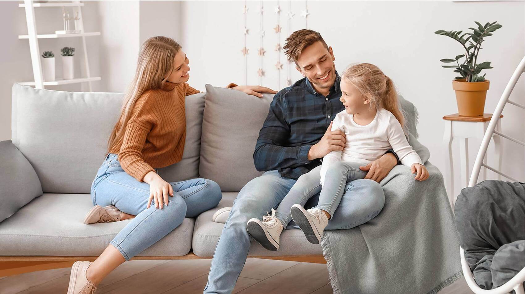 A smiling mother, father, and young daughter sitting together on a grey couch in a bright, modern living room.