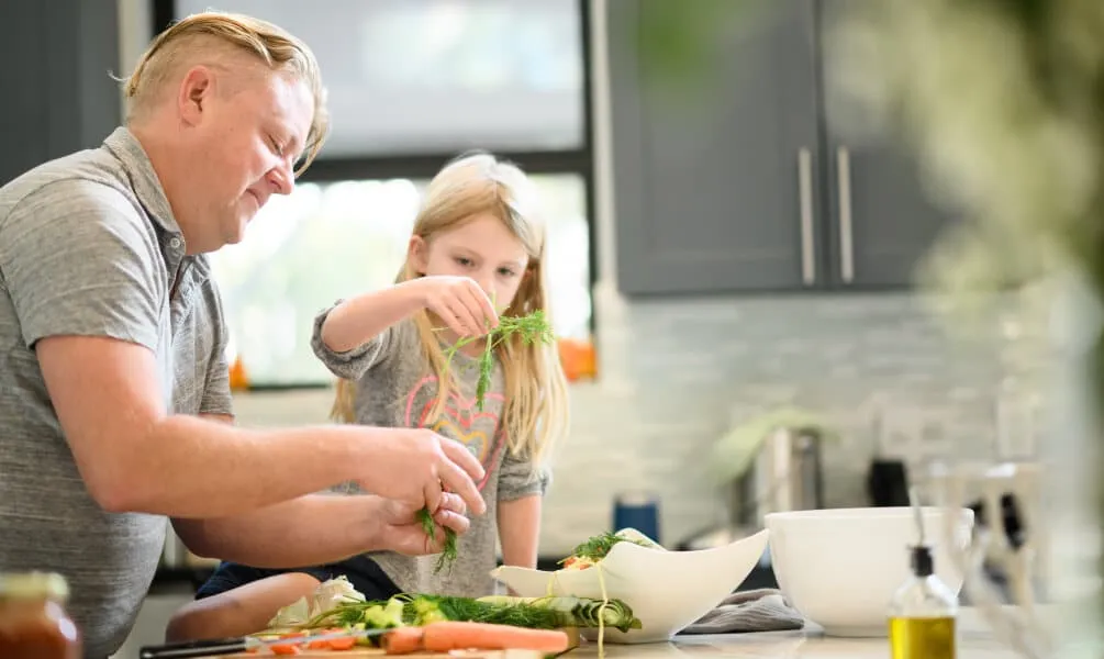 A father and young daughter stand at a kitchen counter together, garnishing a fresh salad with sprigs of dill.