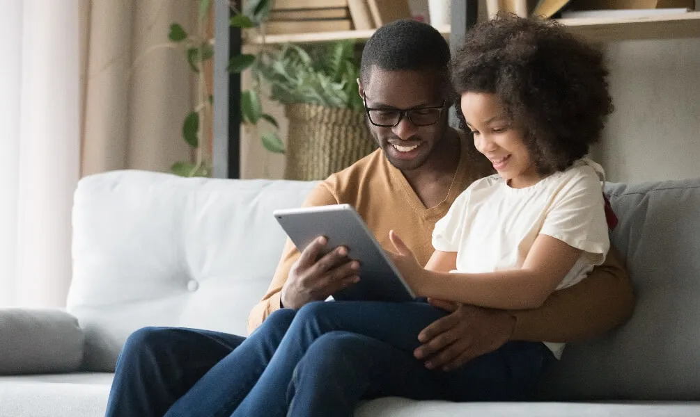 A smiling father and daughter sit on a sofa while looking at a tablet together in a bright living room.