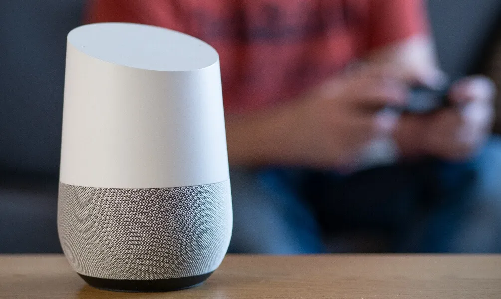 A Google Home smart speaker sits on a wooden surface with a person blurred in the background.