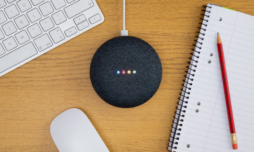 Charcoal Google Home Mini with active lights on a wooden desk next to a keyboard, mouse, and notebook.