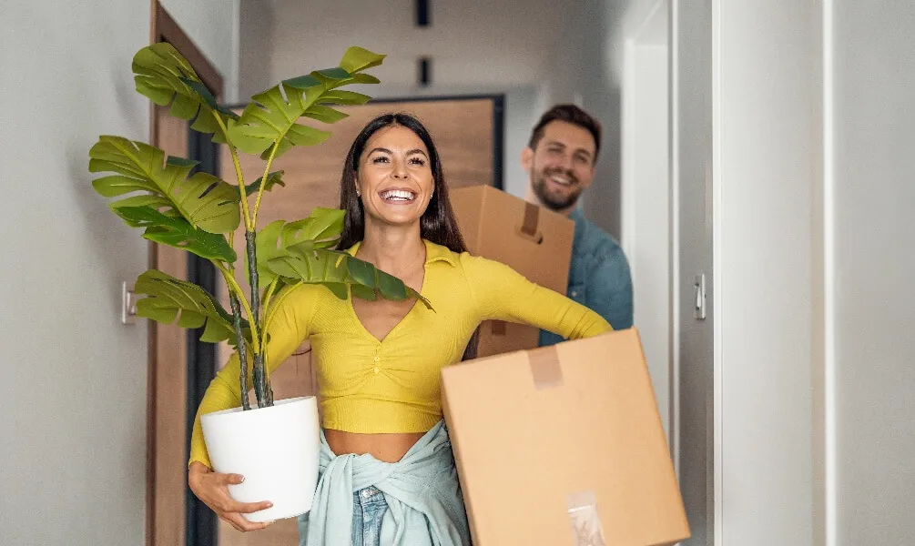 A smiling young woman carries a large potted plant and a cardboard box while moving through a hallway with a man.