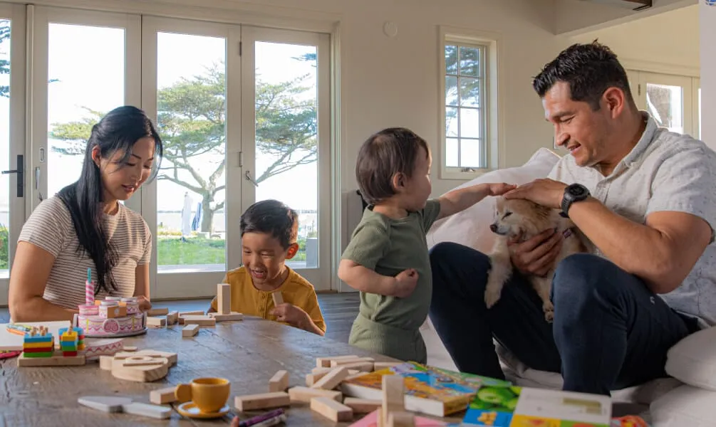 An Asian family of four playing with building blocks on a coffee table in a sunny living room, with the father holding a small dog.