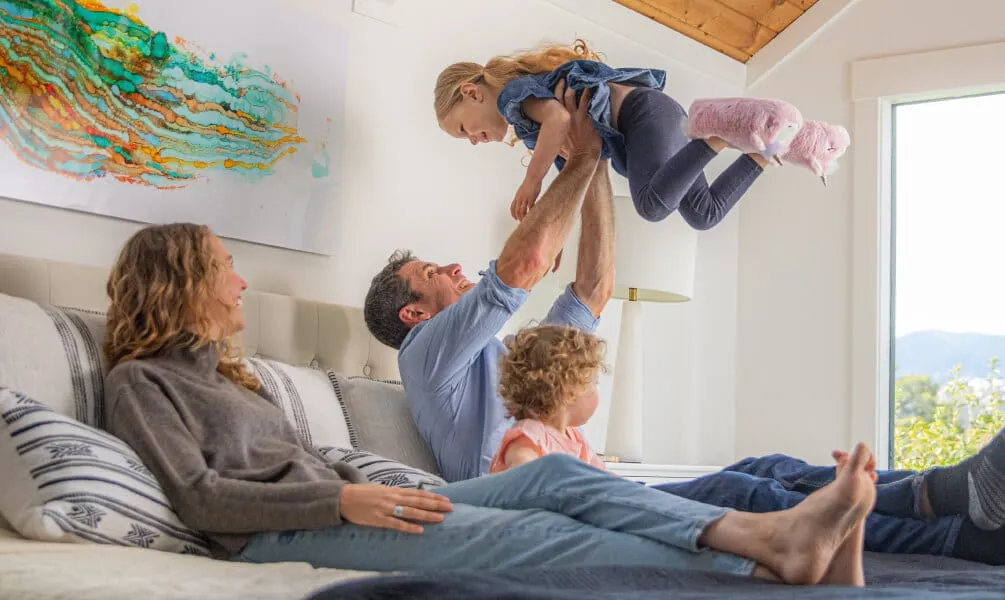 A smiling father holds his young daughter up in the air while lying on a bed with his wife and a younger child.