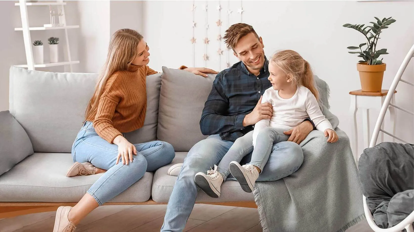 A smiling mother, father, and young daughter sit together on a gray sofa in a bright, modern living room.