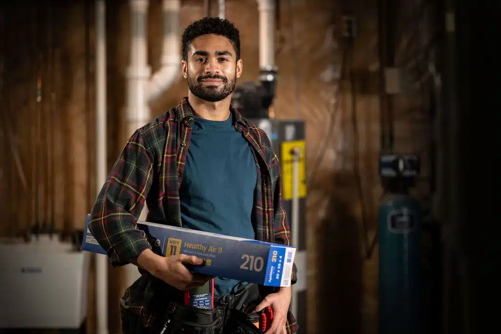 A smiling technician in a plaid shirt stands in a basement holding an AprilAire Healthy Air 210 MERV 11 filter.