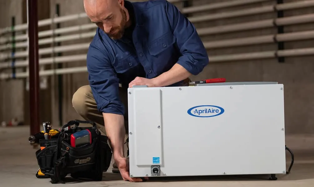 A technician in a blue shirt adjusts the leveling feet of a white AprilAire dehumidifier on a concrete basement floor