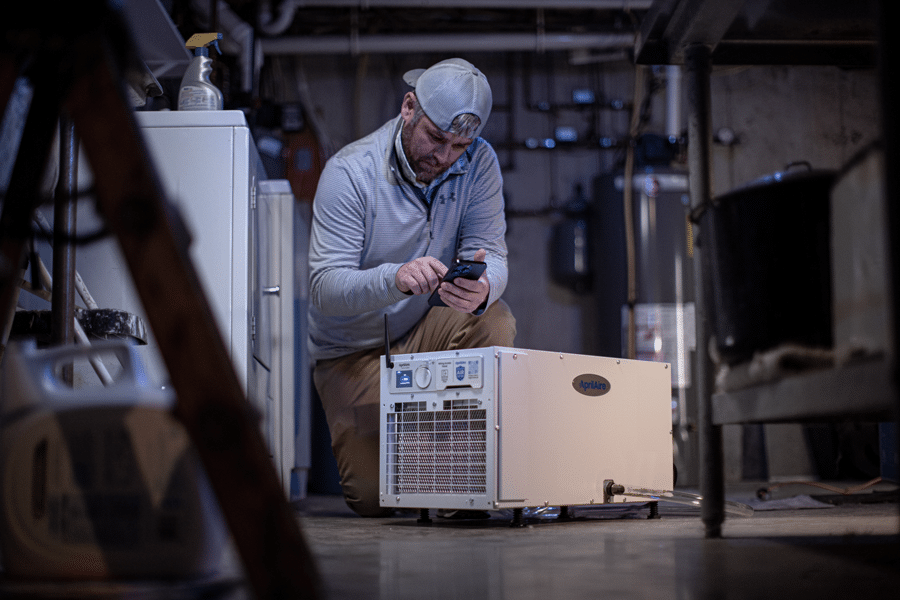 A technician uses a smartphone to configure a white AprilAire dehumidifier unit in a residential basement.