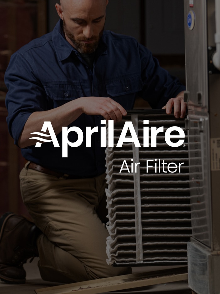 Technician kneeling while sliding a large pleated AprilAire air filter into a residential HVAC furnace system.