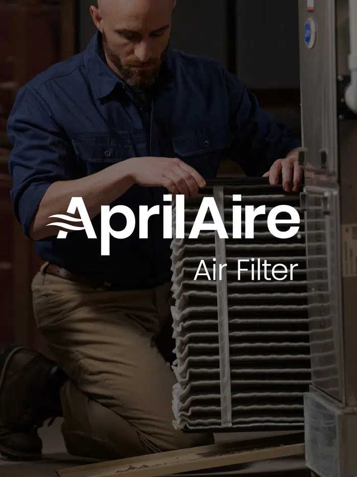 Technician kneeling while sliding a large pleated AprilAire air filter into a residential HVAC furnace system.