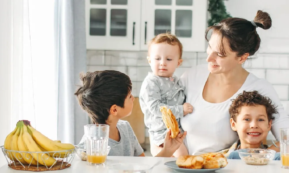 A mother holds a toddler while her two sons eat breakfast and drink juice at a sunlit kitchen table.