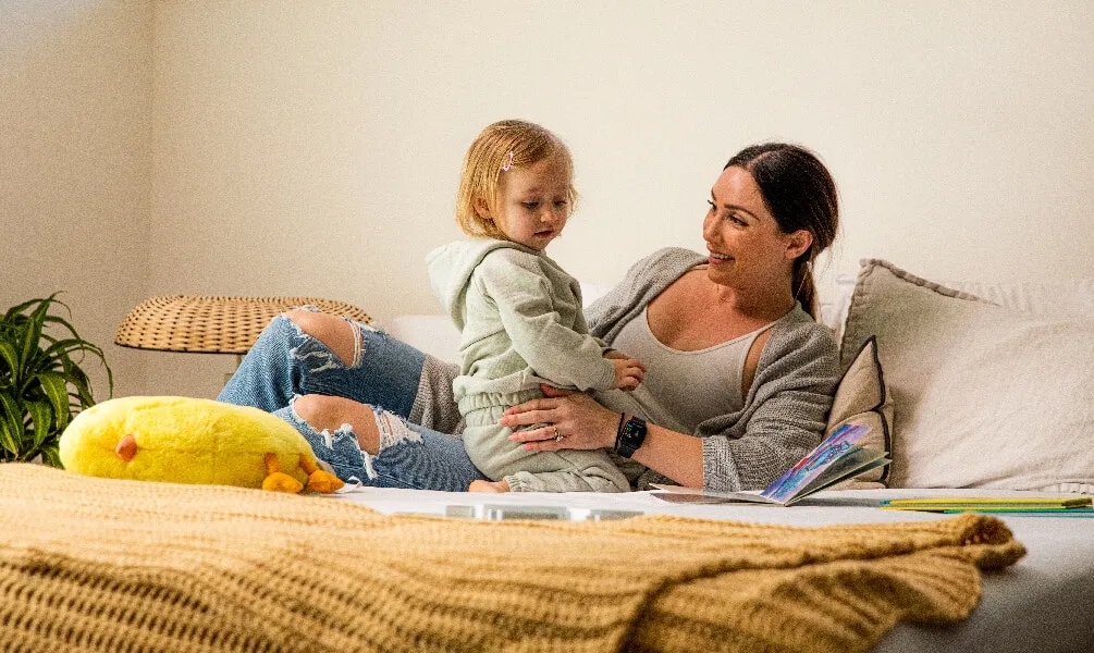 A woman smiles at her young daughter while sitting on a bed with storybooks in a bright, cozy bedroom.