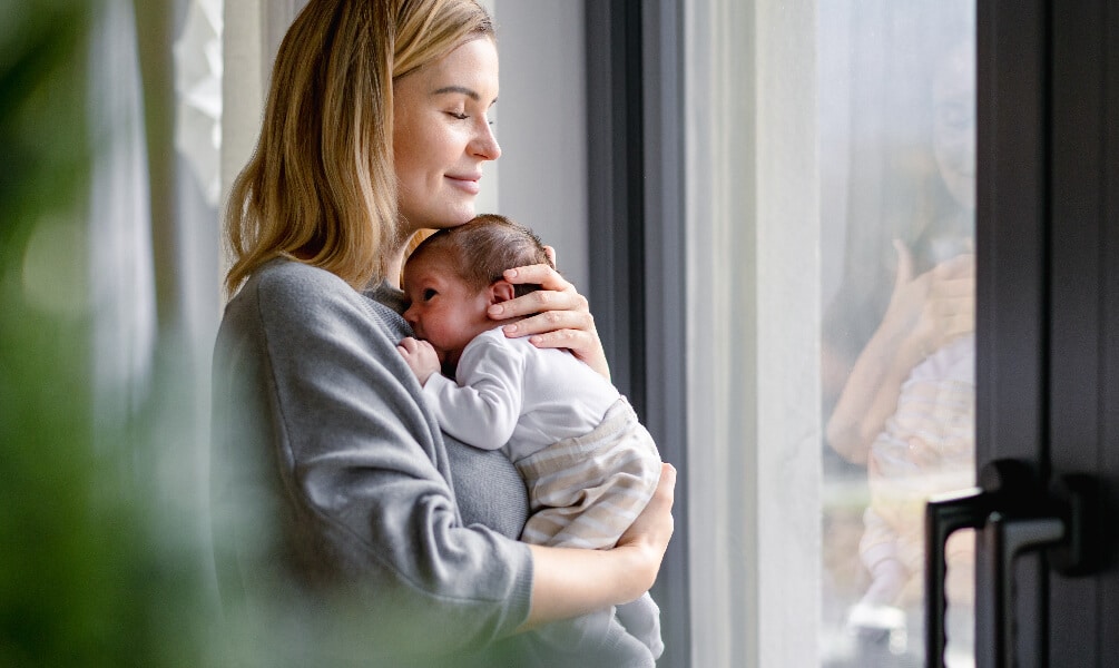 A smiling mother closes her eyes while tenderly holding her newborn baby against her chest near a bright window.