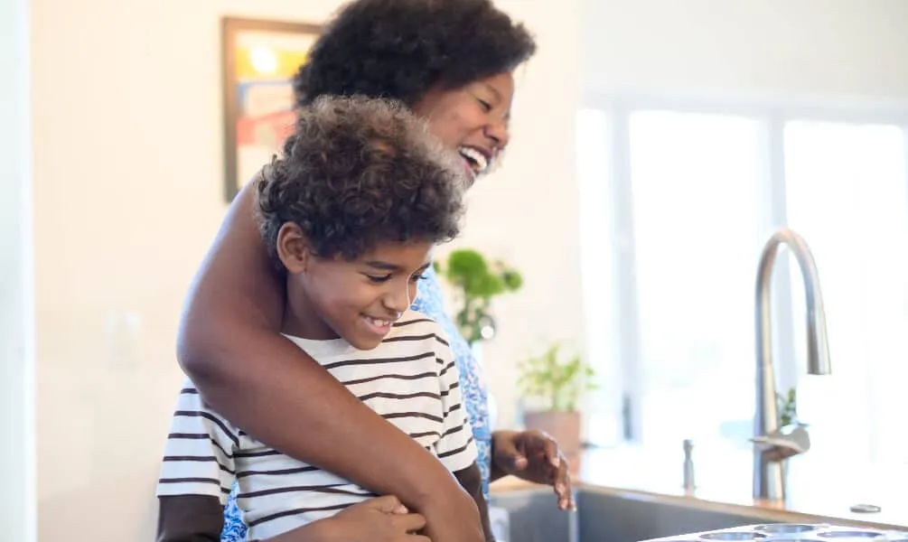A smiling woman embraces her son in a bright, modern kitchen while preparing food together near a sink.