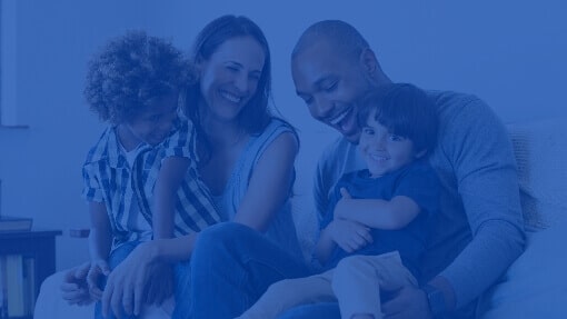 Diverse family of four, including two young children, smiling and laughing together while sitting on a sofa.