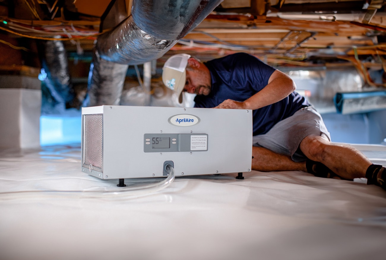 A technician inspects an AprilAire dehumidifier unit inside a white encapsulated residential crawl space.