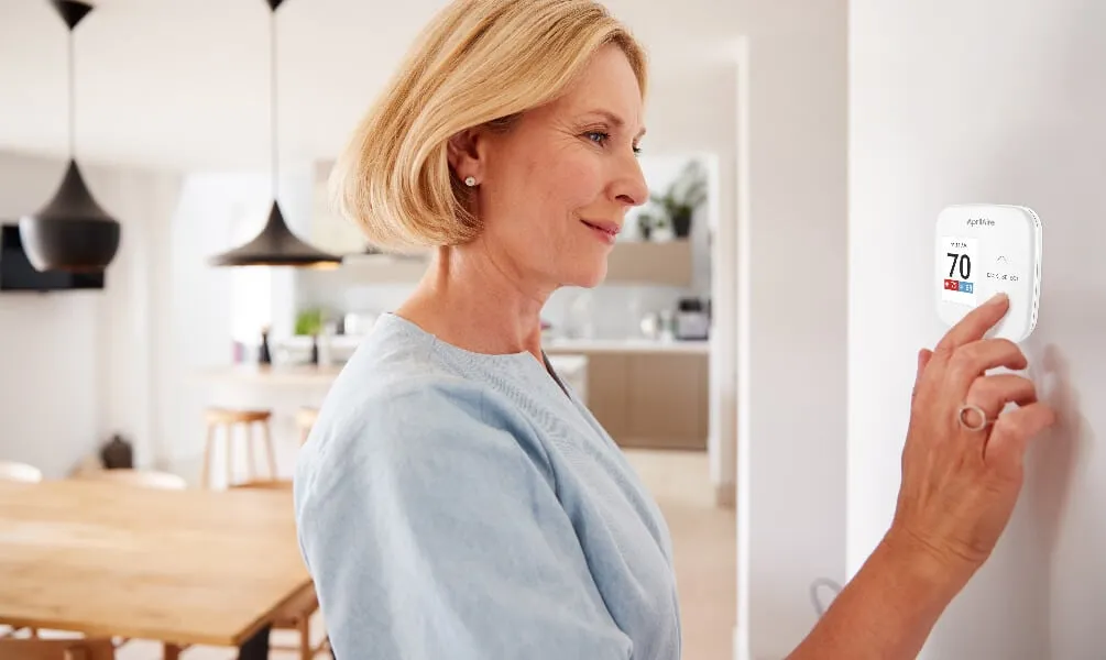 A woman adjusts a digital smart thermostat on a white wall in a modern, sunlit kitchen and dining area.