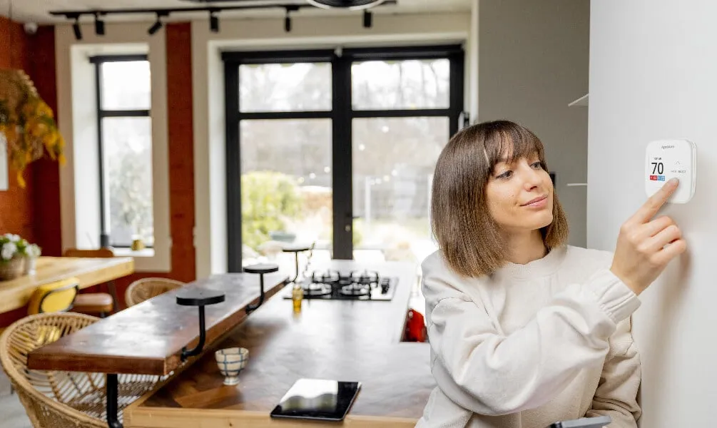 A woman adjusts a digital AprilAire thermostat on a white wall in a modern, sunlit kitchen and dining area.