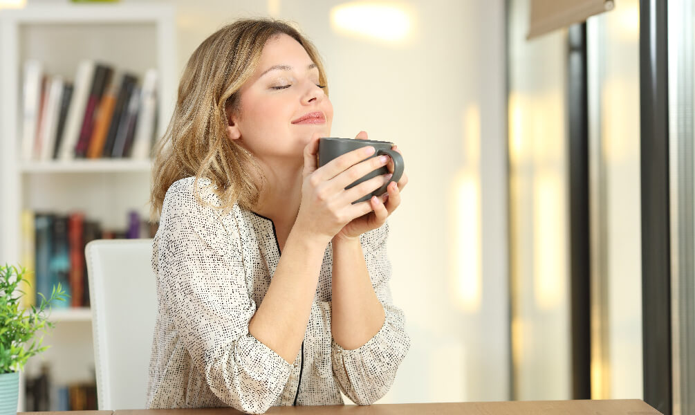 A woman sits at a wooden table with her eyes closed, smiling and holding a mug, enjoying fresh indoor air.