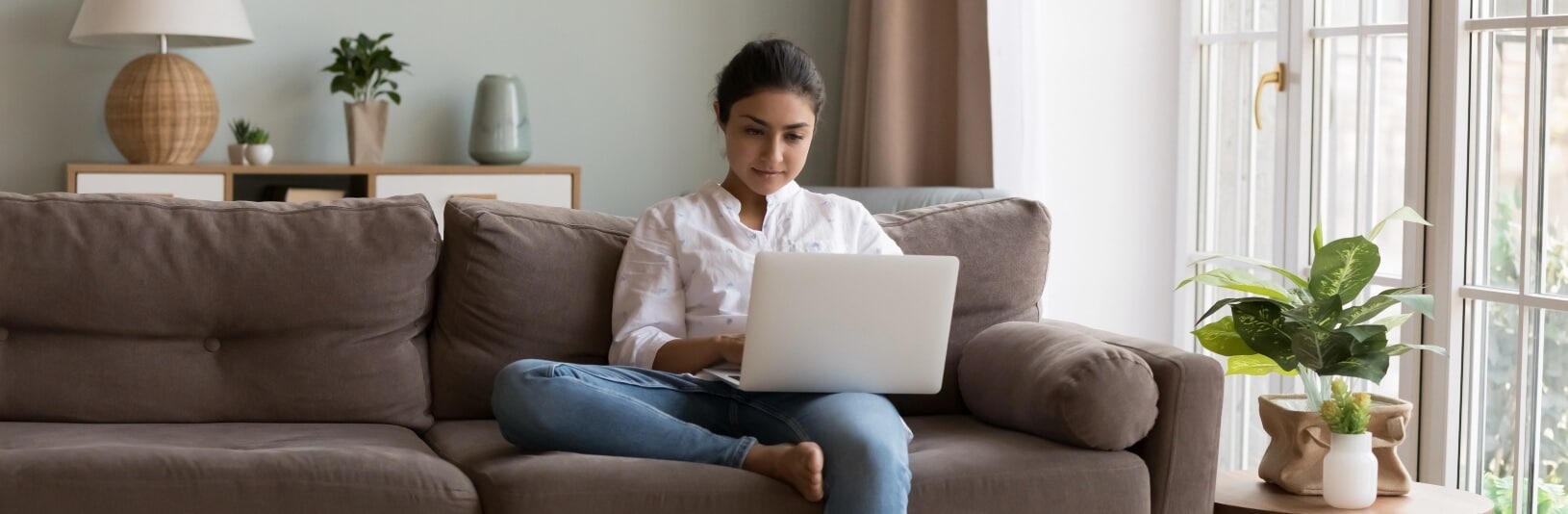A woman sits cross-legged on a brown sofa, focused on her laptop in a bright, modern living room with plants.