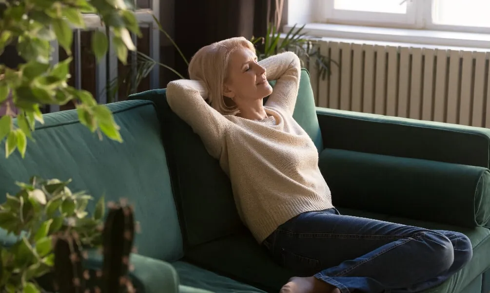 A woman relaxes on a green velvet sofa with her eyes closed and hands behind her head in a bright, plant-filled room.