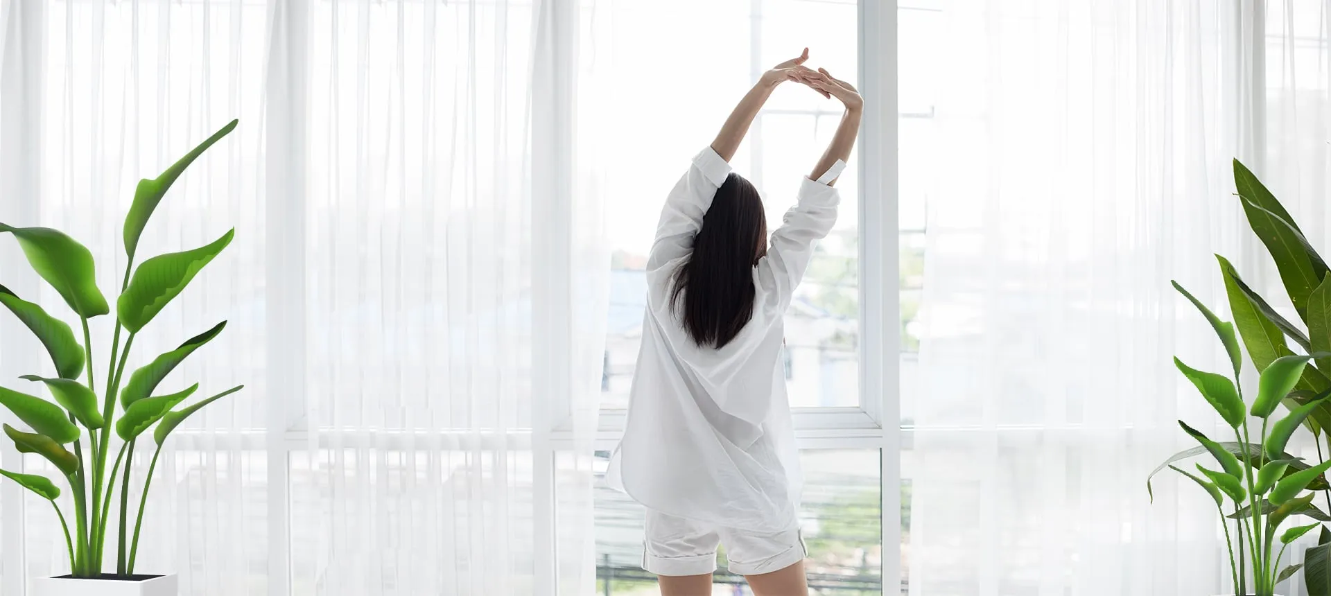 A woman in white clothing stretches her arms upward while standing before large windows with sheer white curtains.