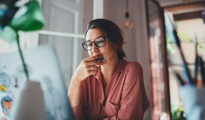 A woman with glasses thoughtfully looks at her laptop screen while holding a pen to her chin in a sunlit room.