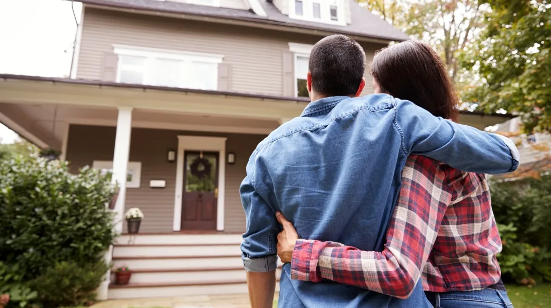 A couple stands with their arms around each other, looking at a two-story house from the front yard.