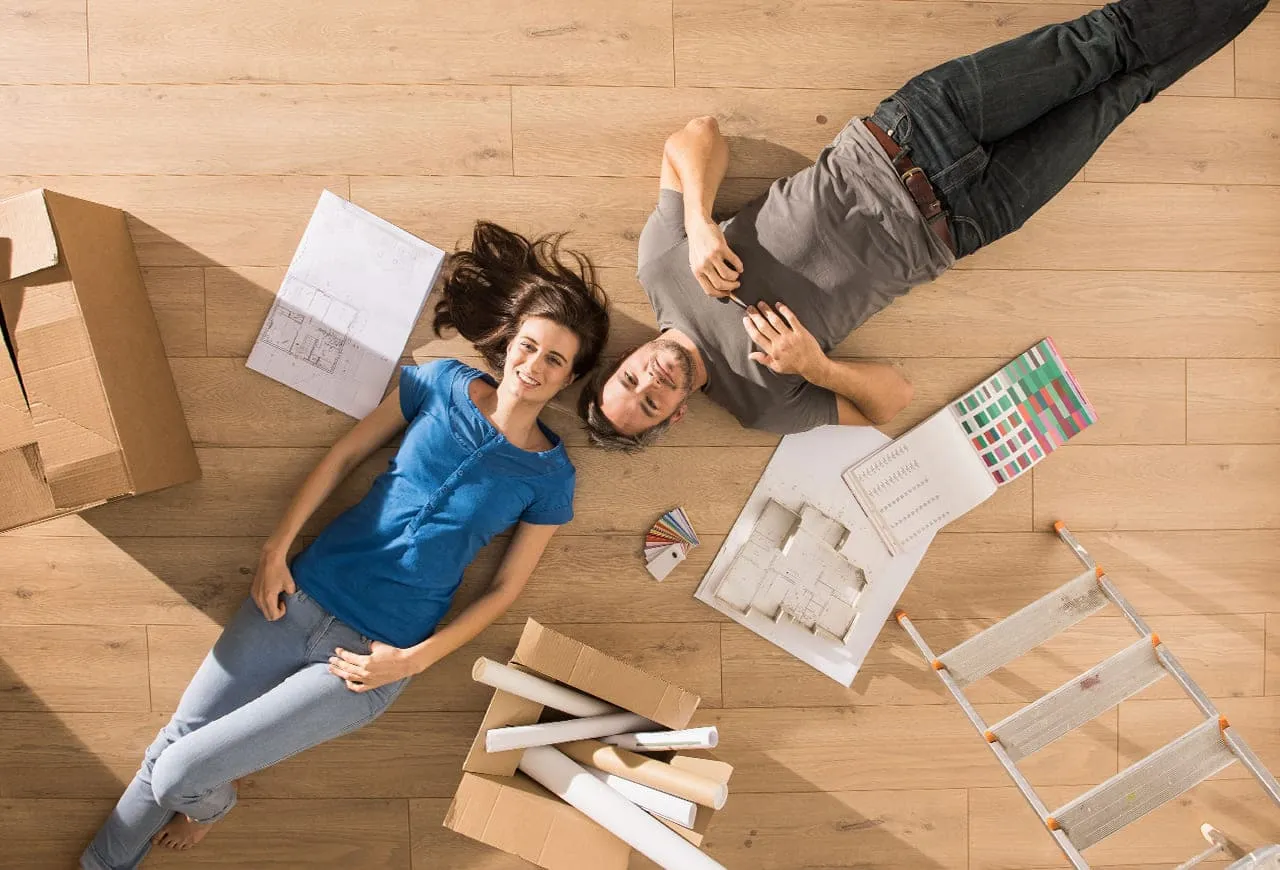 Top-down view of a couple lying on wood floors surrounded by floor plans, color swatches, and moving boxes.