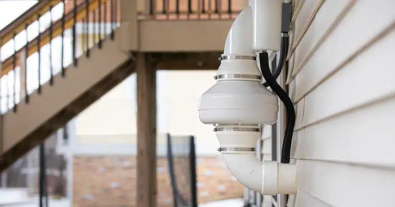 A white radon mitigation fan and PVC piping installed on the beige vinyl siding of a residential home exterior.