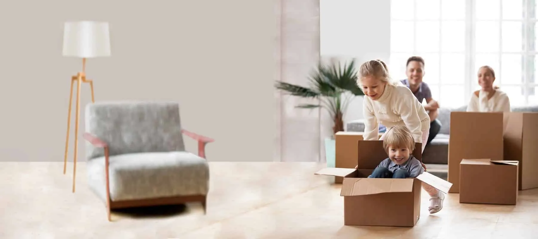 Young girl pushes her brother in a cardboard box while their parents smile in a bright, new living room.