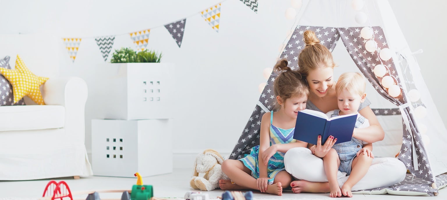 A mother sits in a star-patterned play tent reading a blue book to her two young children in a bright nursery.