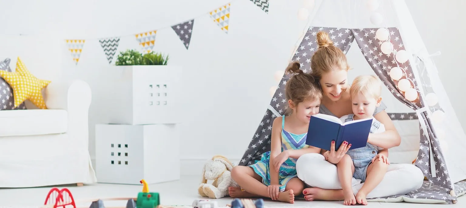A mother sits in a star-patterned play tent reading a blue book to her two young children in a bright nursery.
