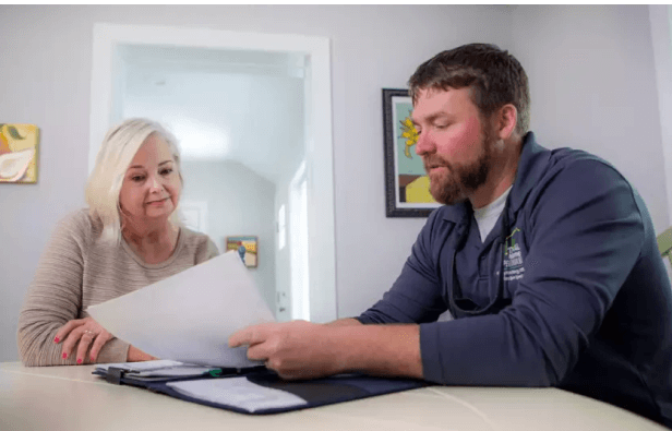 A smiling man wearing an Aprilaire shirt holds a brochure while speaking with a woman near a staircase.