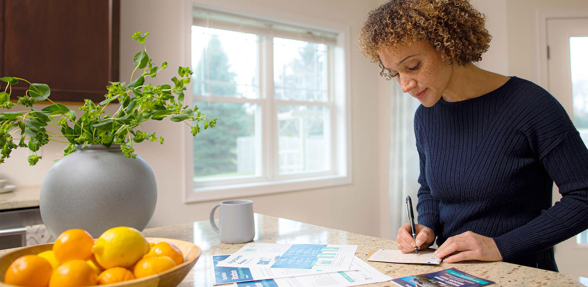 A woman at a kitchen counter fills out paperwork for a short-term radon test kit near a bowl of citrus fruit.