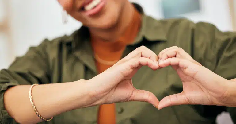 Close-up of a smiling woman in a green shirt forming a heart shape with her hands toward the camera.