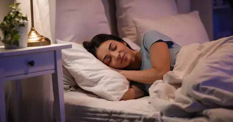 A woman sleeps soundly on white pillows with a soft bedside lamp glowing in a dark, cozy bedroom.