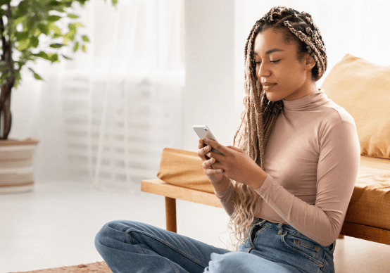 A woman with long braids sits on a rug while looking at her smartphone in a sunlit living room.